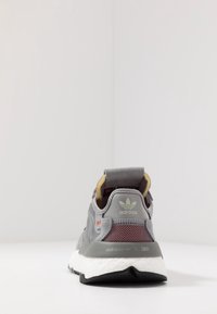 Šedé tenisky s vrchní částí z pružné síťoviny a semiše, černou gumovou podrážkou a bílou mezipodrážkou Boost. Mají zelené logo a červené akcenty.