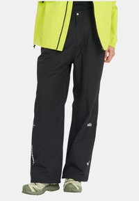 Pantalons imperméables noirs avec une coupe ample, comportant des poches et un logo discret en blanc. Portés avec des chaussures vertes à motifs.