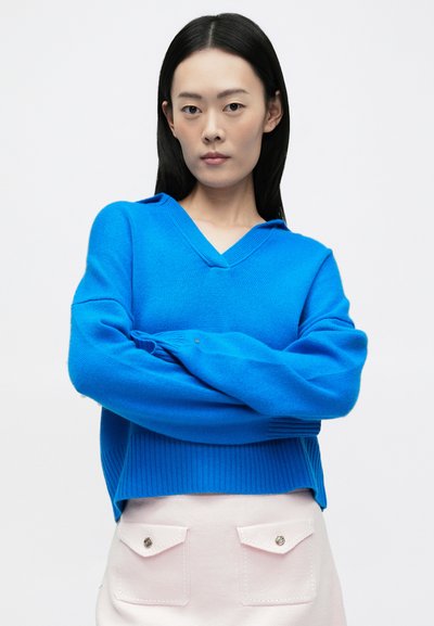 Jasnoniebieski, dziany sweter z dekoltem w kształcie litery V i prążkowanym dołem. Długie rękawy, luźny krój. Połączony z jasnoróżową spódnicą z klapkami na kieszeniach.