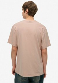 Ung mand med kort brunt hår iført en ensfarvet lysebrun T-shirt med korte ærmer og blå jeans, set bagfra.