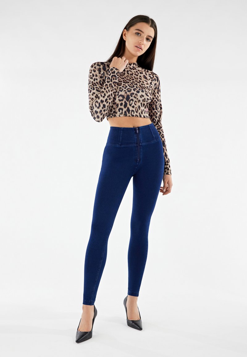Crop top a maniche lunghe con stampa leopardata e collo alto, abbinato a jeans skinny a vita alta blu scuro e décolleté neri a punta.