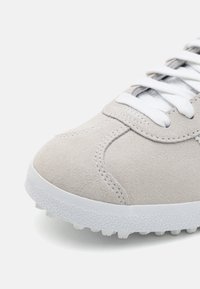 Sneaker din suede gri deschis, cu șireturi albe, guler căptușit și o talpă din cauciuc alb texturată, dotată cu mici crampoane pentru aderență.