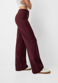 Kvinna som bär högmidjade, vida burgundyröda byxor med vita sneakers och en kort vit topp, stående mot en plain vit bakgrund.