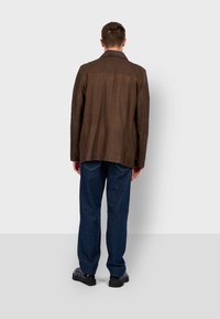 Chaqueta de cuero marrón con un diseño estructurado, que presenta un cuello, corte recto y botones laterales. Combinada con pantalones de mezclilla azules.