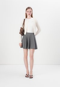 Cardigan blanc léger et moelleux sur un haut blanc ajusté, associé à une jupe évasée grise et des tongs noires, accessoirisé avec un sac marron.