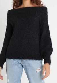 Czarny sweter z odkrytymi ramionami i luźnymi rękawami, wykonany z miękkiego, puszystego materiału. Połączony z jasnoniebieskimi, podartymi dżinsami.