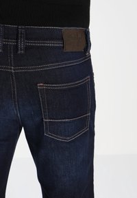 Mörkblå denimjeans med kontrasterande sömmar och en läderlogg i midjan, med en synlig bakficka.
