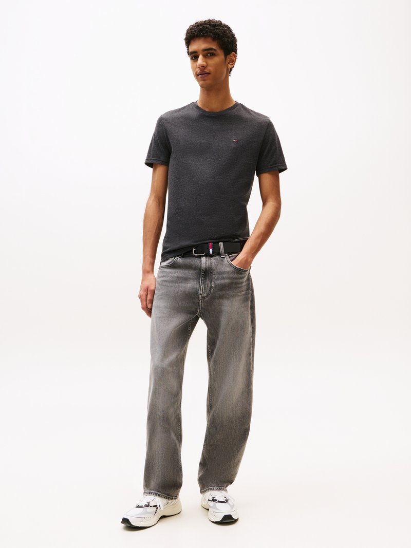 Jeune homme debout avec une main dans la poche, portant un t-shirt gris foncé, un jean gris délavé, une ceinture noire et des baskets blanches sur un fond uni.
