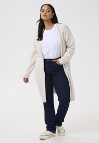 Cardigan lungo beige con lavorazione a maglia testurizzata, indossato su una t-shirt bianca e jeans blu scuro. Completato con sneakers bianche, design minimalista.