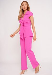 Woman in bright pink short-sleeve tie-front blouse and matching pants, walking in beige high-heeled sandals against plain background.