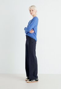 Blå strikkegenser, litt oversized, kombinert med mørke vide bukser og svarte joggesko med hvite striper og beige detaljer.