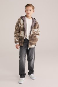 Barna és krémszínű polárdzseki kapucnival, elöl cipzárral és texturált mintával. Fehér póló fölé viselve, szürke farmerrel és fehér cipővel párosítva.