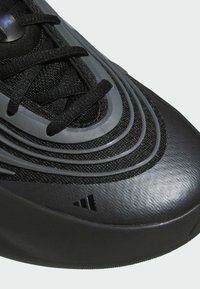 Chaussure de sport noire avec une tige en tissu texturé, des superpositions en plastique gris et des lacets noirs. Présente des détails nervurés et un logo distinct.