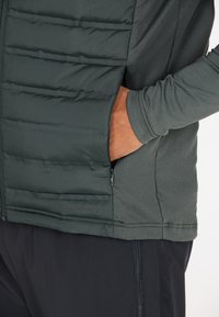 Mørkegrøn isoleret jakke med en glat overflade, der har quiltet polstring på forsiden og en lynlåslomme på siden.