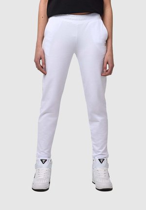Personne portant un pantalon de jogging blanc slim-fit et des baskets blanches montantes, debout devant un fond gris clair.