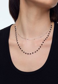 Dwie naszyjniki warstwowe z srebrnymi łańcuszkami. Niższy naszyjnik ma czarne koraliki; górny naszyjnik ma delikatny, prosty wzór.