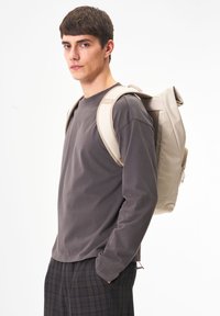 Hellebeiger Rucksack mit gepolsterten Trägern, strukturiertem Design und einem Reißverschlussfach. Getragen von einer Person in einem grauen Langarmshirt und karierten Hosen.