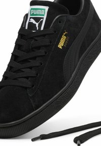 Sneaker in suede nera con suola in gomma testurizzata, lacci neri piatti e un'etichetta con il logo Puma verde e bianco. Brandizzazione dorata Puma sul lato.