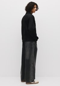 Giacca in suede nera con colletto alto, abbinata a jeans grigi scuri a gamba larga e sneakers color crema, con una vestibilità rilassata.
