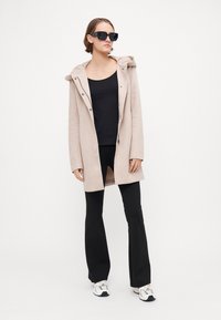 Hellbeige Kapuzenmantel mit Knopfleiste, schwarzes Tanktop, schwarze Schlaghose und weiße Sneaker mit silbernen Akzenten.