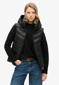 Gilet matelassé noir avec capuche, comportant des sections matelassées, porté par-dessus un pull en maille noir. Associé à un jean bleu et une ceinture marron.