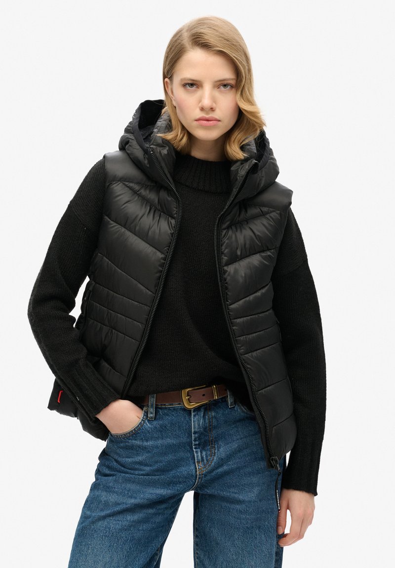 Gilet matelassé noir avec capuche, comportant des sections matelassées, porté par-dessus un pull en maille noir. Associé à un jean bleu et une ceinture marron.