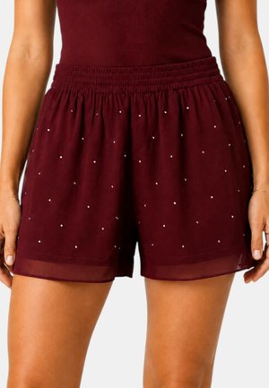 Donna che indossa pantaloncini bordeaux a vita alta con piccoli pois bianchi e un top aderente abbinato, in piedi con le mani rilassate lungo i fianchi.