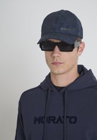 Gorra de marineros hecha de tela suave con visera curva, que presenta el logo "ANTONY MORATO". Combinada con gafas de sol cuadradas negras y una sudadera navy.