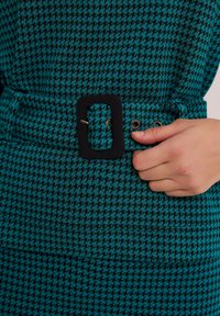 Tkanina w wzór houndstooth w kolorze turkusowym i czarnym ma prostokątną czarną klamrę, zapiętą metalowym zatrzaskiem i oczkami.