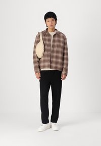 Giacca camicia a quadri marrone, maglione strutturato crema, pantaloni neri tapered, sneakers bianche; il modello tiene una borsa beige su una spalla.