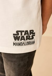 Tricou alb din bumbac cu un logo imprimat negru care spune "STAR WARS THE MANDALORIAN" situat aproape de tiv. Fit casual cu mâneci scurte.