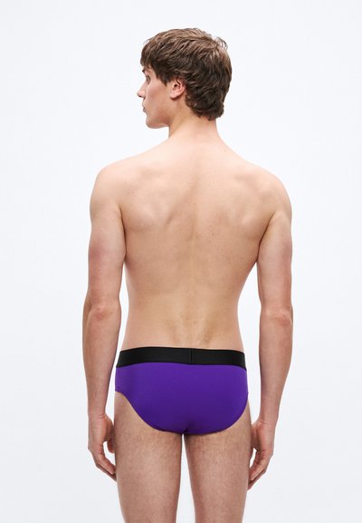 Lila úszónadrág fekete derékpánttal, sima textúrával és testhezálló kialakítással, hátulnézetben egy férfi modellen.