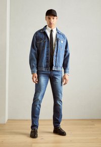 Ung mann iført denimjakke og jeans, hvit skjorte, svart slips, svart caps og svarte sko som står på tregulv foran en ensfarget vegg.