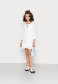 Vestido branco até ao joelho com decote quadrado e mangas longas; corpete texturizado; combinado com uma mala azul clara e sandálias chunky.