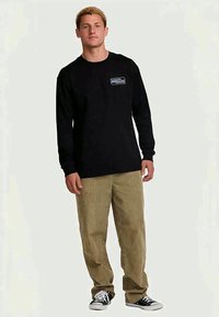 Camisa preta de mangas longas com um pequeno logotipo no peito, combinada com calças de veludo cotelê em tom bege claro e sapatilhas pretas. Design simples e casual.