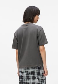 Camiseta de algodón gris, de mangas cortas, con cuello redondo. Presenta una parte posterior lisa y una pequeña etiqueta de la marca en el dobladillo. Combinada con una falda de cuadros.