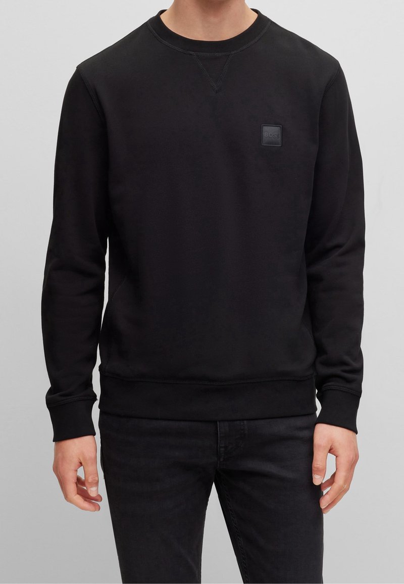Homme portant un sweat-shirt noir à manches longues et col rond avec un petit patch carré sur la poitrine gauche, associé à un pantalon noir.