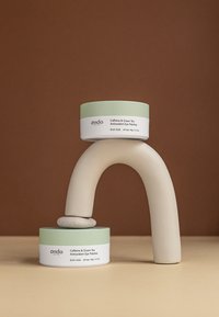 Due contenitori color crema con coperchi verde menta, etichettati "Patch oculari antiossidanti al caffeina e tè verde", disposti su una scultura curva beige.