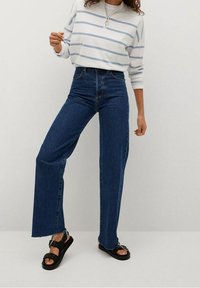 Jeans in denim blu a vita alta con gamba dritta e orlo sfrangiato, indossati con una maglietta a maniche lunghe a strisce bianche e azzurre e sandali neri.