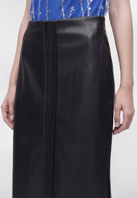 Femme portant une jupe en cuir noir taille haute avec un détail de couture verticale, associée à un top bleu orné de sequins argentés.