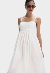 Witte zondress met een smock-buste, dunne straps en een vloeiende rok. De stof heeft subtiele textuurdetails en een lichtgewicht ontwerp.