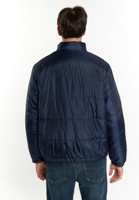 Giacca imbottita blu scuro con una texture liscia e lucida, dotata di colletto alto e polsini elasticizzati, indossata sopra dei jeans blu.