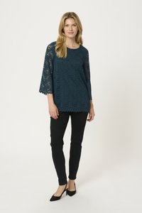 Top in pizzo turchese con maniche a tre quarti e orlo scallopato, abbinato a pantaloni neri slim-fit e tacchi neri. Texture liscia e motivo intricato.
