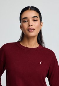 Burgunderfarbener Pullover aus weichem Stoff mit klassischem Rundhalsausschnitt und kleinem gesticktem Figuren-Logo auf der linken Brust.