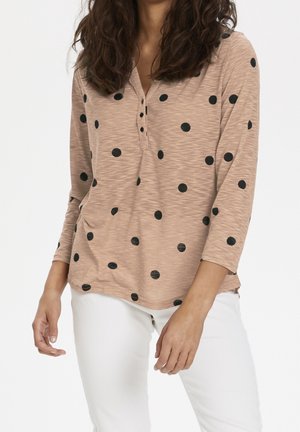 Chemise à manches longues marron clair avec des pois noirs, dotée d'un col henley et d'un tissu doux et texturé. Portée avec un pantalon blanc.