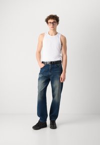 Débardeur sans manches blanc associé à un jean en denim bleu, avec une coupe décontractée et un léger délavage. Une ceinture noire et des chaussures de sport noires complètent le look.