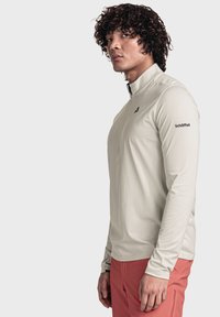 Helles beige Langarmshirt mit Viertelreißverschluss aus glattem Material, mit schwarzem Logo auf dem linken Ärmel und taillierter Passform.