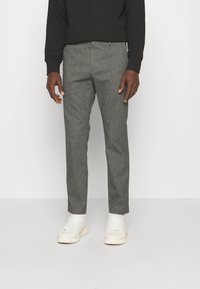 Homme portant un pantalon gris cintré, des baskets blanches et une chemise noire à manches longues, debout devant un fond blanc uni.