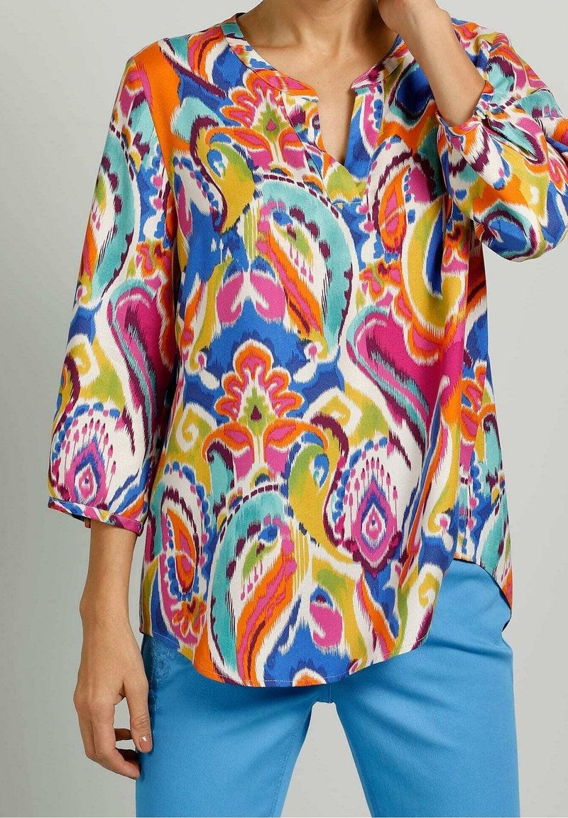 Femme portant un chemisier coloré à motif cachemire avec des manches trois-quarts et un pantalon bleu, debout avec une main près du visage.