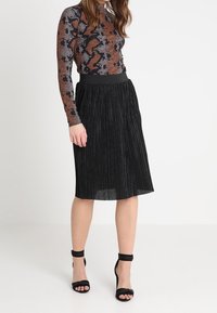 Svart plisserad kjol med hög midja, matchad med en mönstrad långärmad topp. Skorna har svarta ankelremmar med klack.